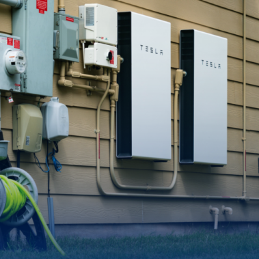 Most Popular Solar Battery Options: A Tesla Powerwall Battery is shown on the side of a house next to electrical boxes.