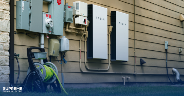 Most Popular Solar Battery Options: A Tesla Powerwall Battery is shown on the side of a house next to electrical boxes.