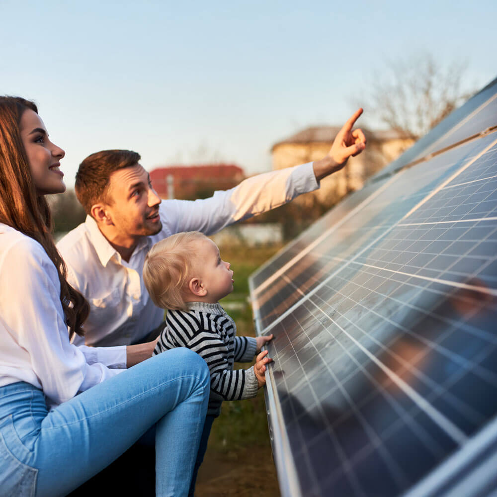 A family—two adults and a small child—looks at and interacts with a ground-mounted solar panel array outdoors.