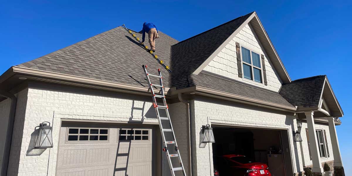 A worker in a blue shirt climbs a ladder to the asphalt-shingle roof of a white suburban home on a clear, sunny day.