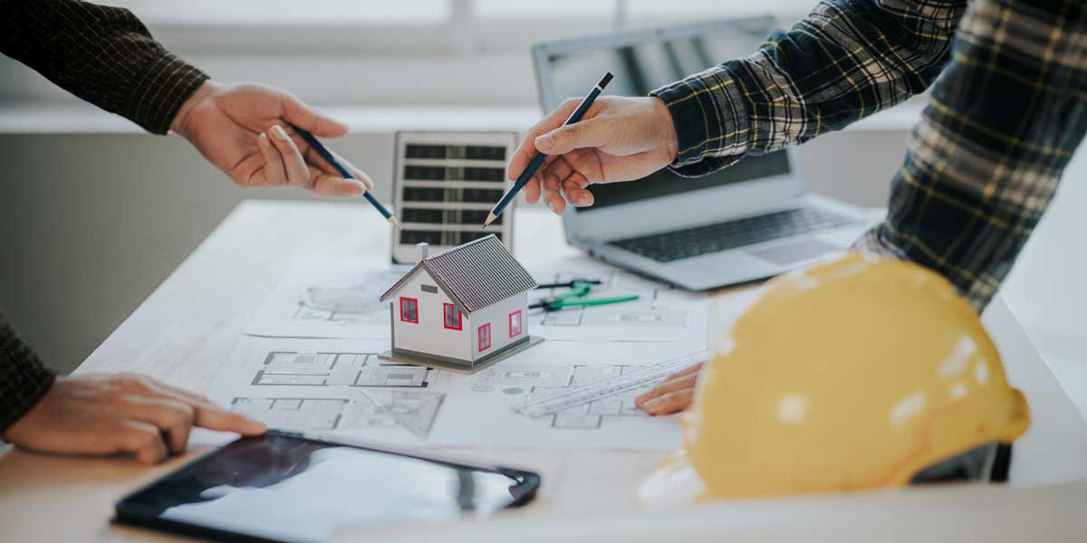Two people point pencils at a miniature house model placed on architectural blueprints, with a small solar panel and yellow hard hat nearby.