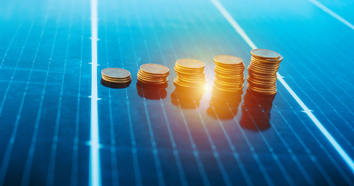 Stacked gold coins of increasing height arranged in a rising bar-chart pattern on top of a blue solar panel.