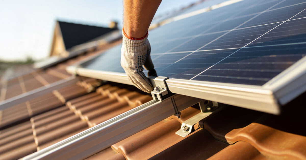 A gloved hand securing a solar panel to mounting rails on a terracotta tile roof.